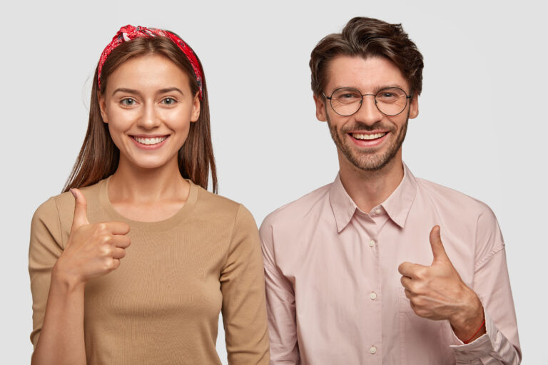 Horizontal shot of pleased satisfied girlfriend and boyfriend raise thumbs, have toothy smiles, stand closely to each other against white background, being in good mood